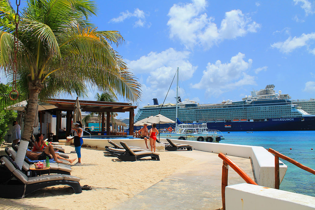 small elevated beach shoreline with access to the sea of turquoise waters with a large cruise line ship in the background