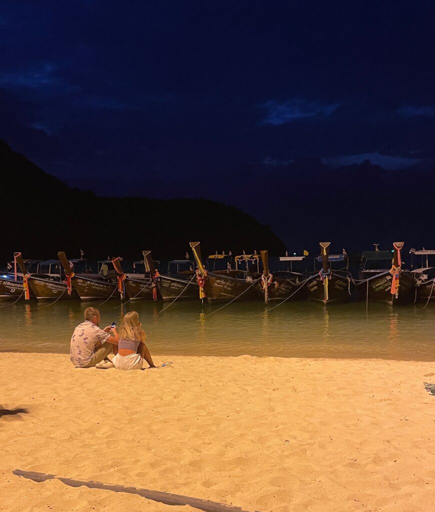 several longtail boats docked on the beach in Phi Phi, a couple sits under the lights on the beach