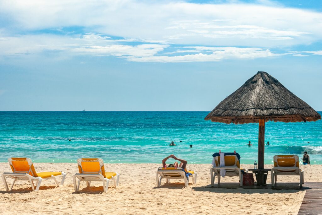 beach lounge chairs under a palapa on a beautiful beach with clear skies and various shades of turquoise waters in Cancun Mexico