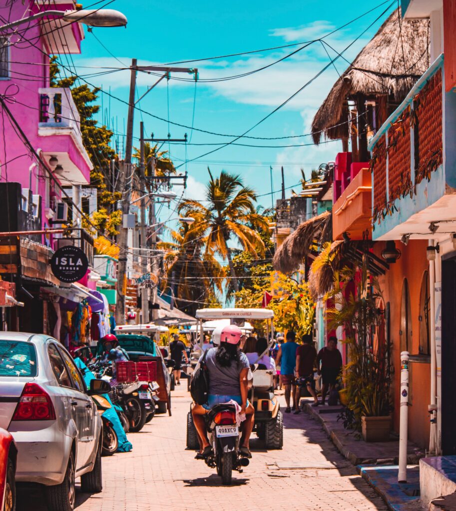 A woman is sitting on the back of a motorcycle with bright coloured buildings on the sides of the streets and palm trees/ clear skies in the distance