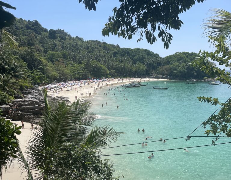 views of the entire Freedom Beach shoreline showing several tourist on the beach, beautiful turquoise waters and dense green forest surrounding the sand in Phuket, Thailand