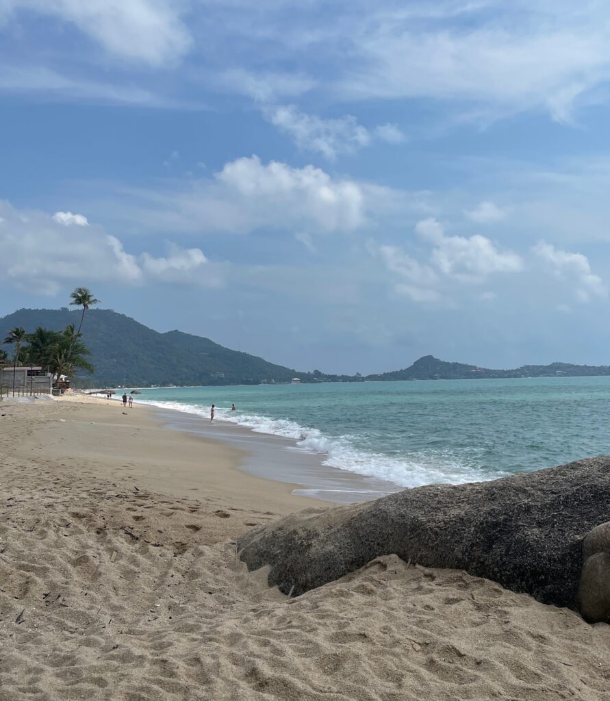 long beach shoreline showing mountains, palm trees and clear skies in the distance at Lamai Beach in Koh Samui, Thailand