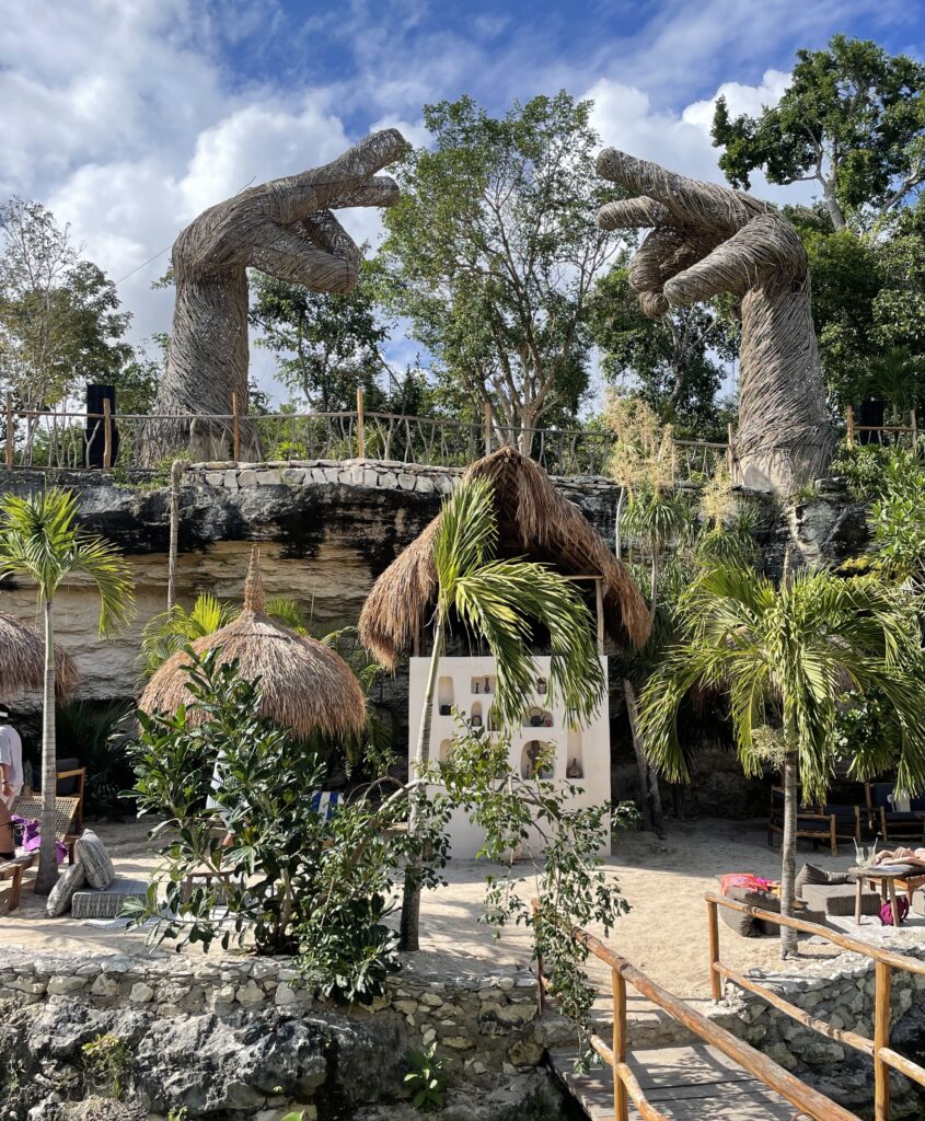 photo from an inside view of a cenote in Casa Tortuga Tulum where a beach club is, view of a DJ booth, clear skies and two large hand sculptures