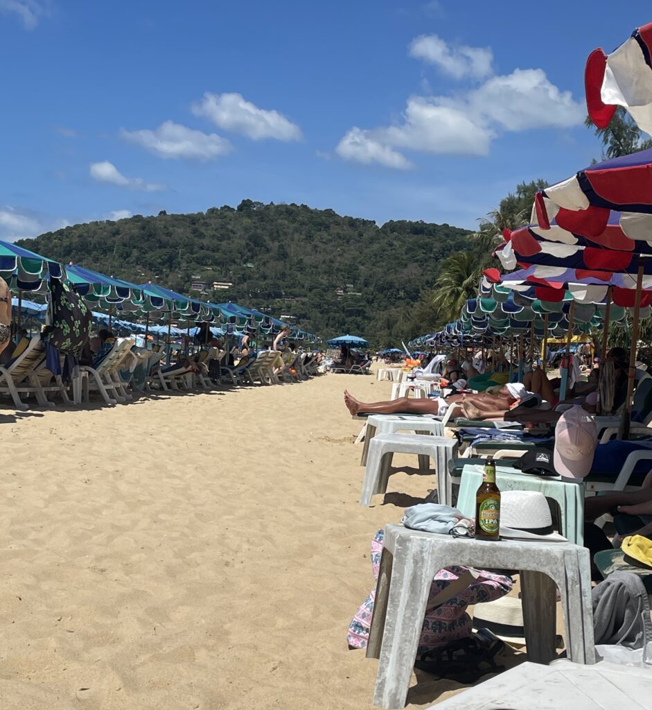 several tourists sunbathing under umbrellas and clear skies, mountainous range in the distance at Karon Beach Phuket