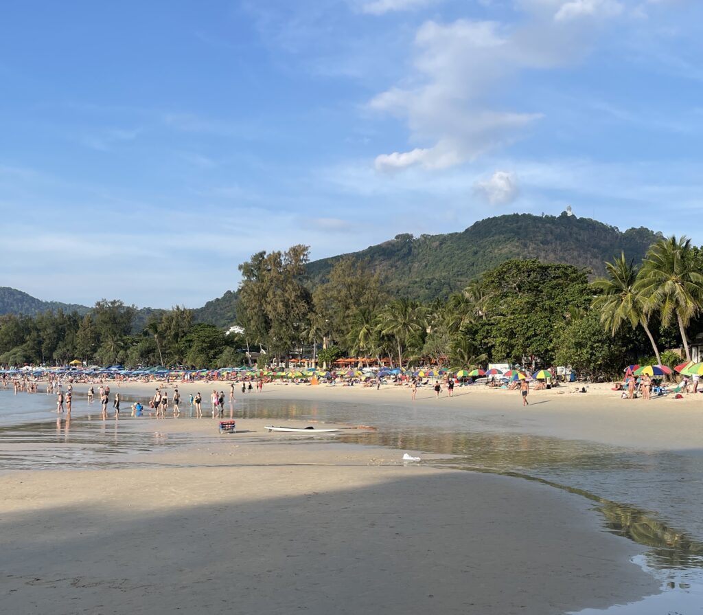 a busy extensive beach shoreline full of people at Kata Beach and a mountain range in the background in Phuket, Thailand, is Phuket expensive?