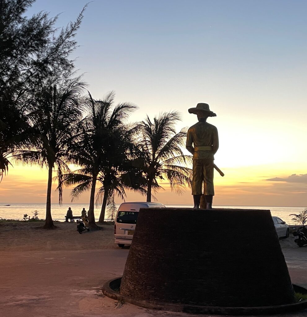 a stunning sunset setting in with several palm trees and a small statue of a man with a hat in Karon Park Phuket Thailand