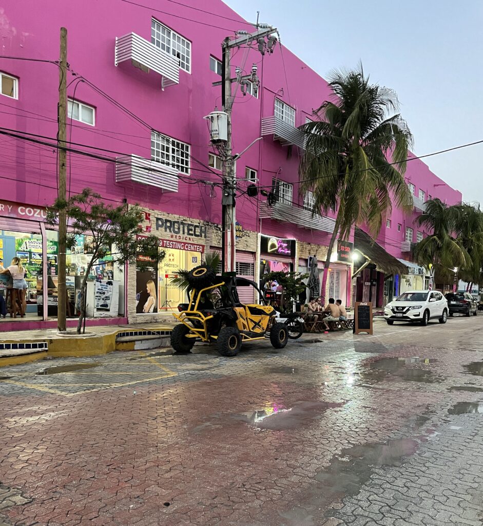 a dune buggy parked on the side of the road with a pink building in the background and palm trees along the road in Playa Del Carmen, Mexico