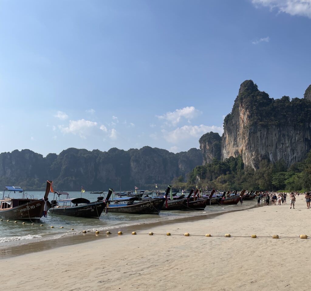 several tourists walking by the long tail boat taxis in Railay Beach / how to get from Phuket to Railay Beach / Is Railay Beach worth visiting