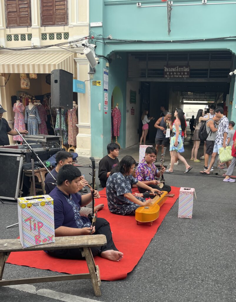 three Thai children sitting on the road playing instruments at the Old Phuket Sunday Market