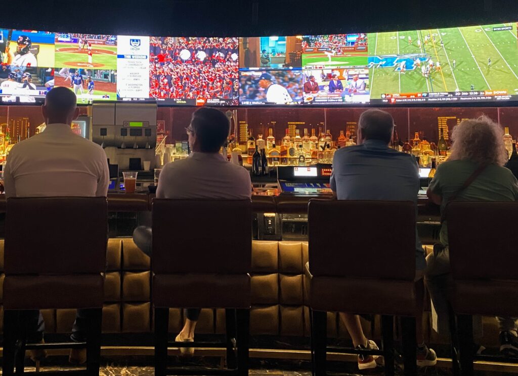 a few guys sitting at the bar in front of large tvs displaying several sports
