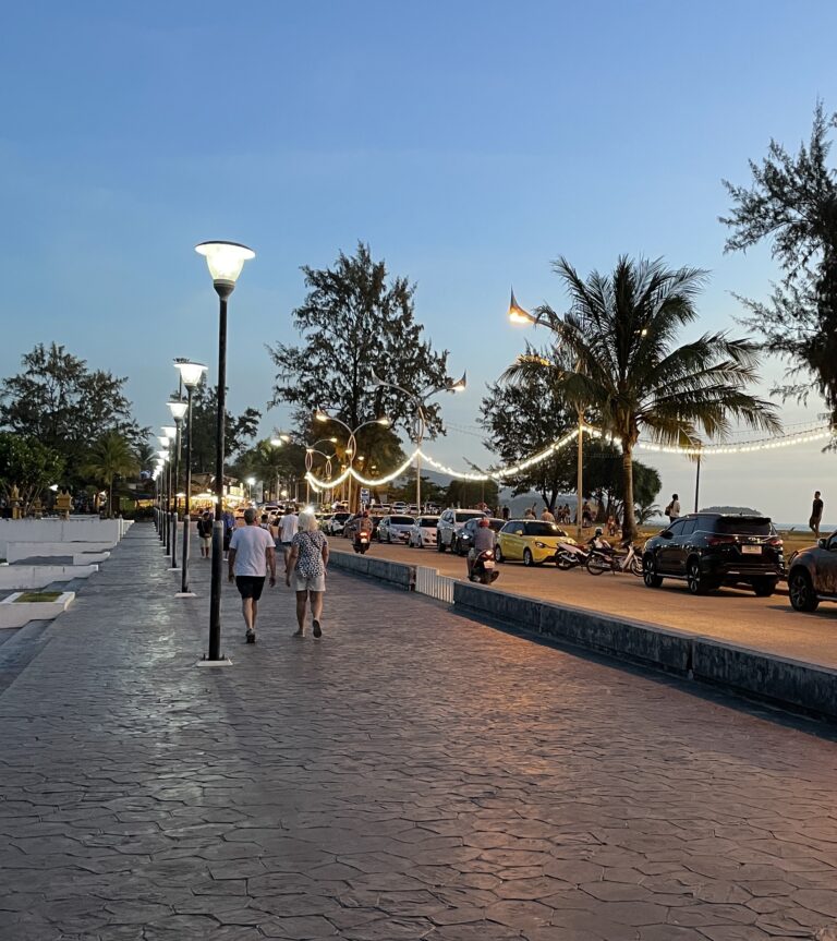 old couple walking along the Karon Beach boardwalk in Phuket at sunset amongst clear skies and palm trees / best things to do in Karon Beach