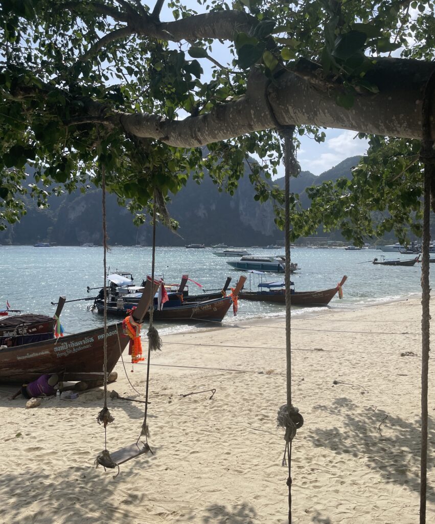 a wooden swing hanging on a tree with many Thai longtail boats on the shoreline on a beach in Koh Phi Phi in Krabi
