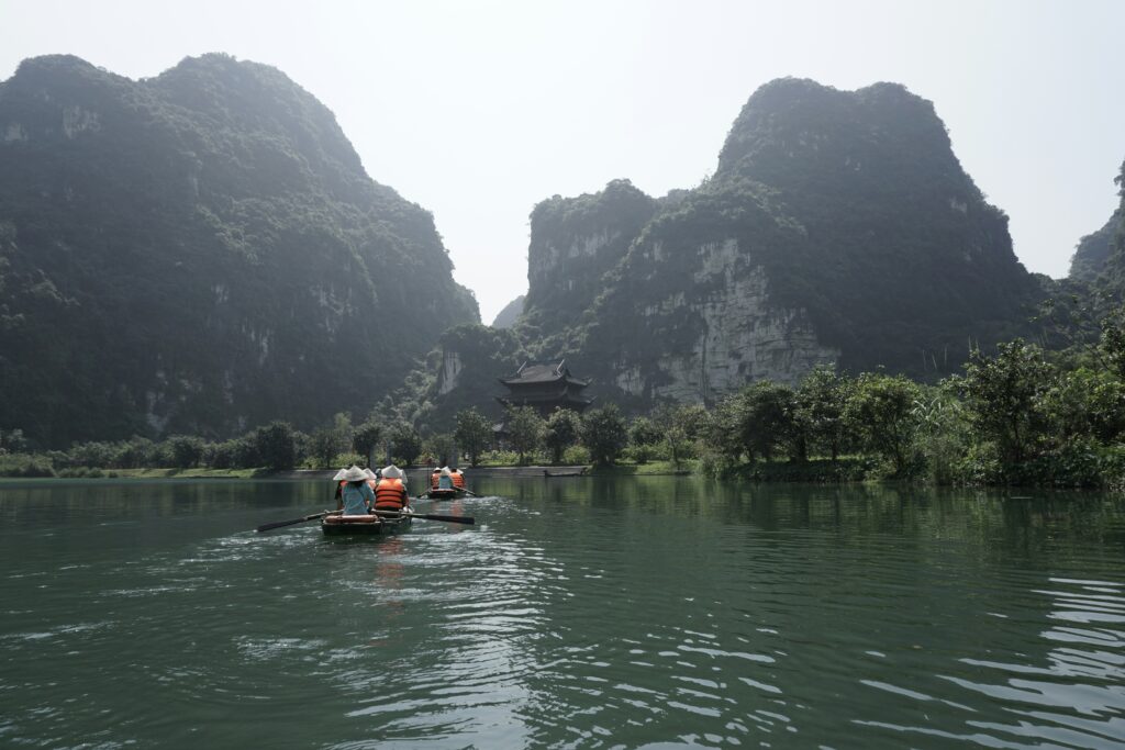 locals riding through the water with towering limestones and temples in the background, in Ninh Binh, Vietnam