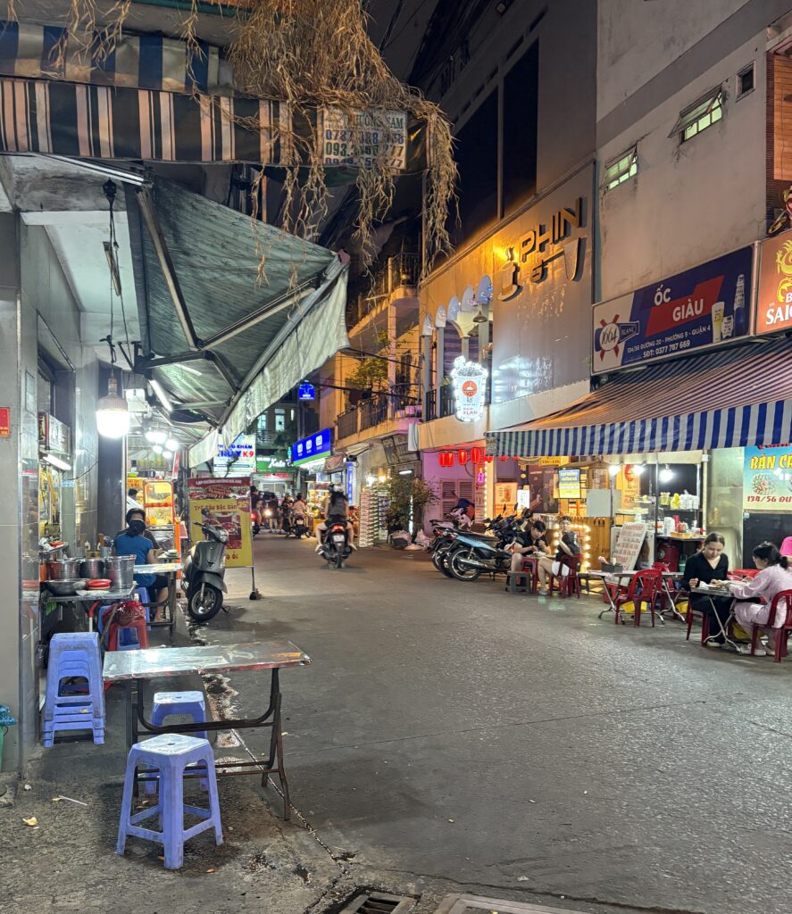 Wide alleyway in Ho Chi Minh City lined with food stalls and bustling with people dining under warm street lights