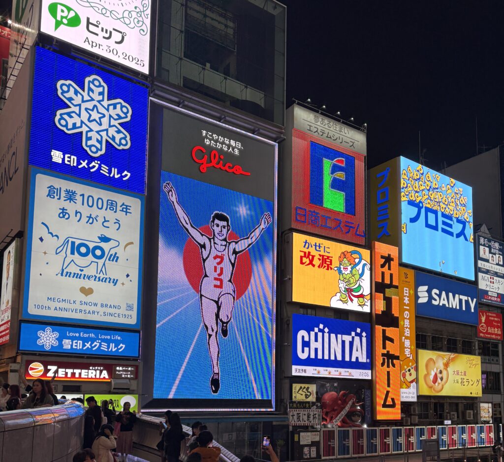 The iconic Glico running man sign illuminated at night in Dotonbori, surrounded by vibrant neon advertisements and city crowds in Osaka.