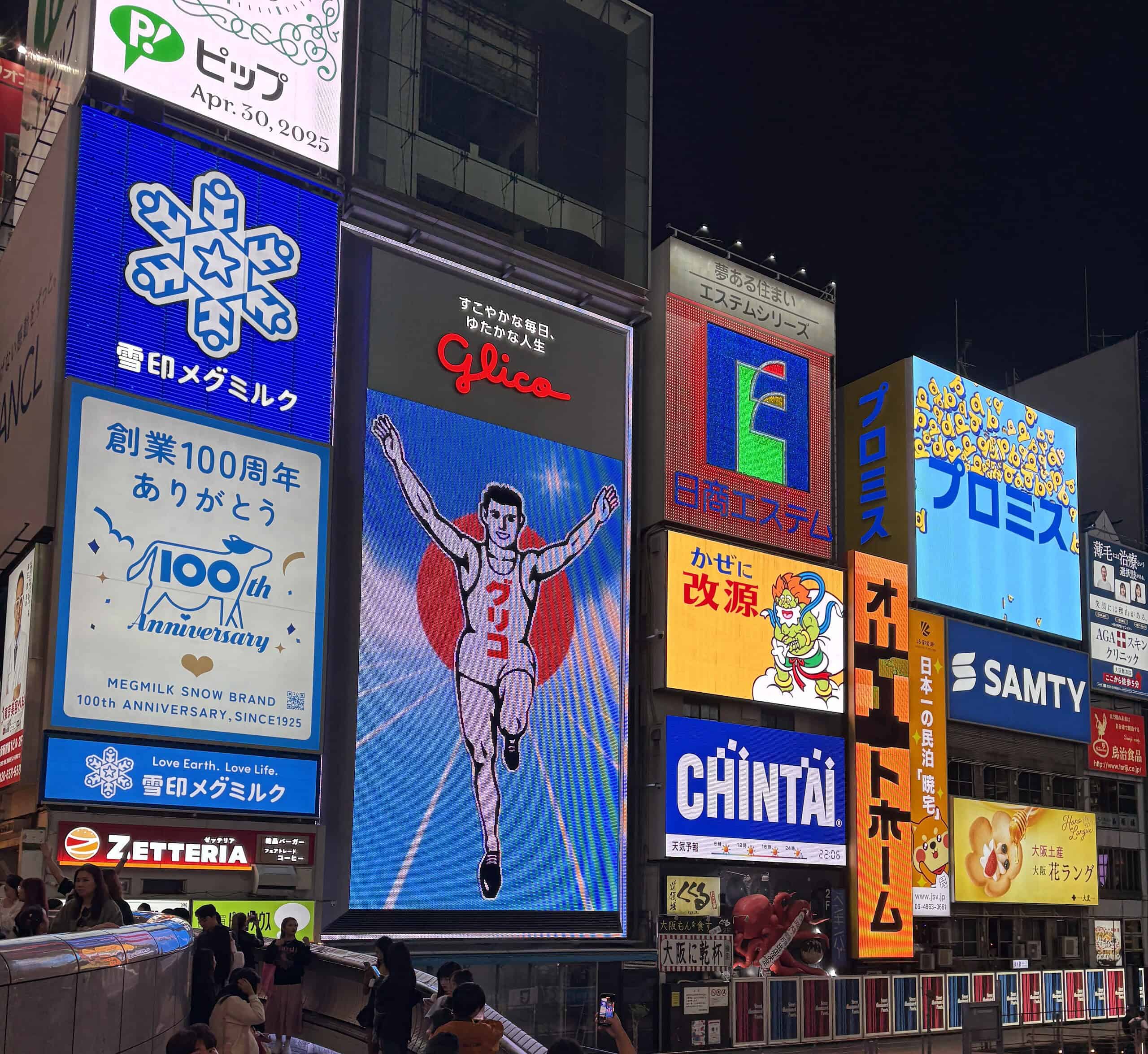 The iconic Glico running man sign illuminated at night in Dotonbori, surrounded by vibrant neon advertisements and city crowds in Osaka.
