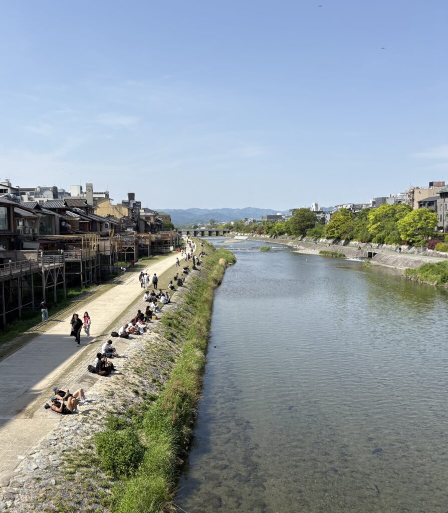 Locals and tourists relaxing by the Kamogawa River in Kyoto with distant mountains on a sunny day