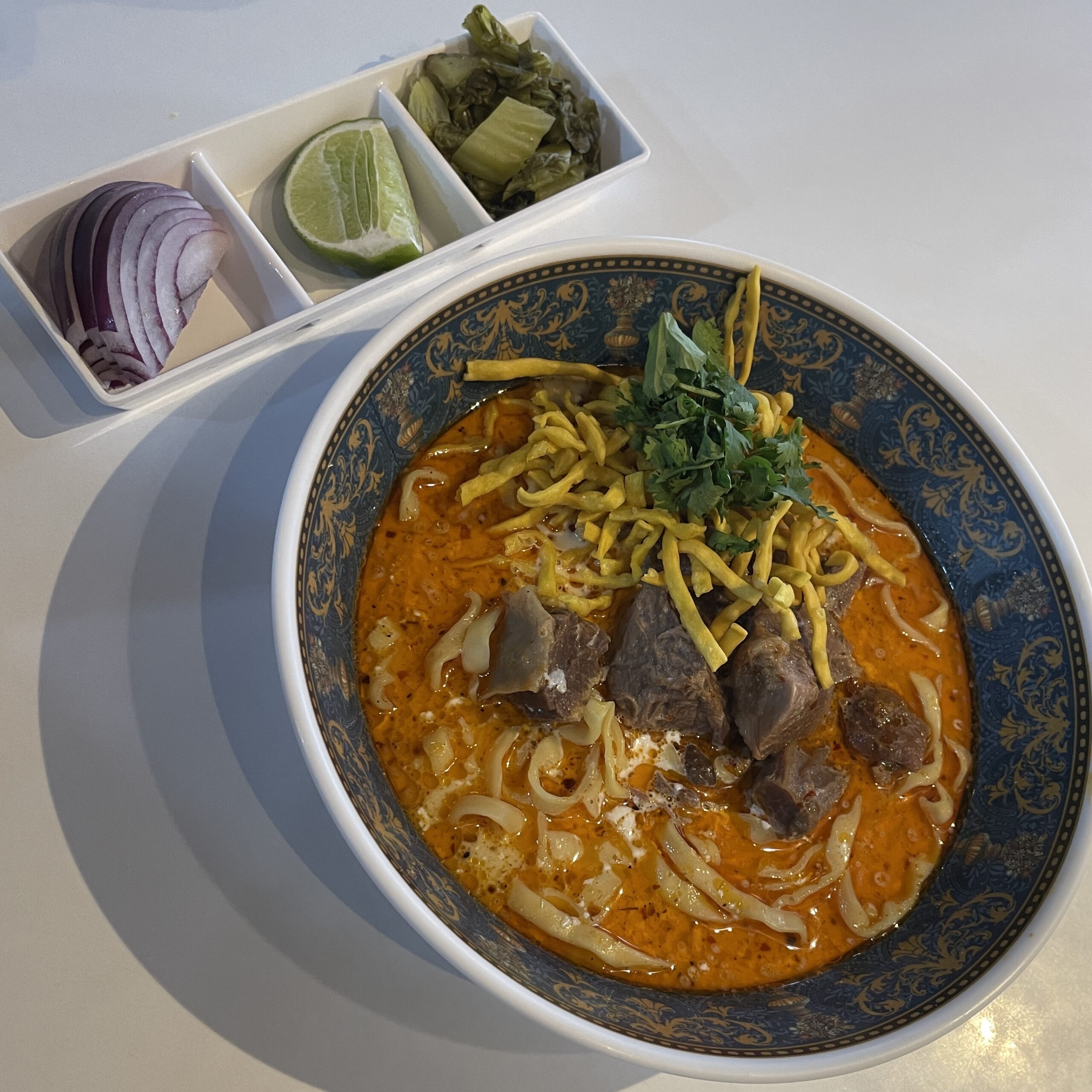 A vibrant bowl of Khao Soi with rich red coconut curry broth, served with three classic condiments—onions, pickled greens, and lime—in Chiang Mai, Thailand.