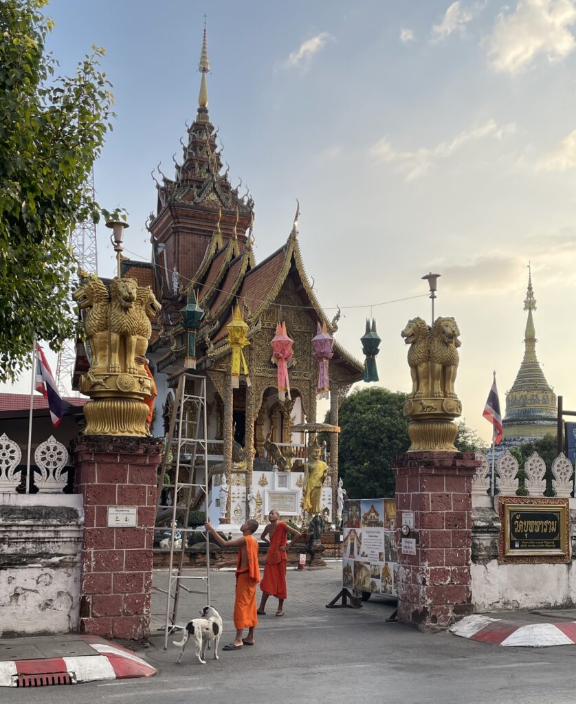 Two monks with a dog beside them setting up decorations at a temple in the Chang Moi area of Chiang Mai during the evening hours.