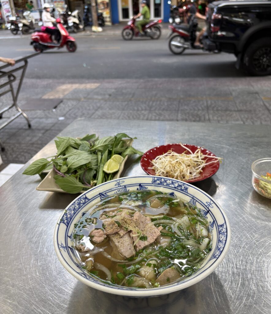 Close-up of a steaming bowl of pho served on a metal table in Ho Chi Minh City with fresh toppings and scooters passing by in the background