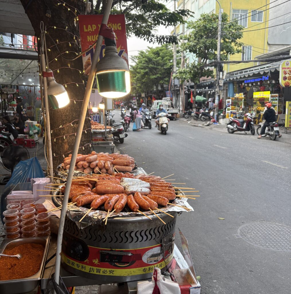 Street vendor grilling pork sausages on hot stone with scooters passing by near the Dragon Bridge in Da Nang city