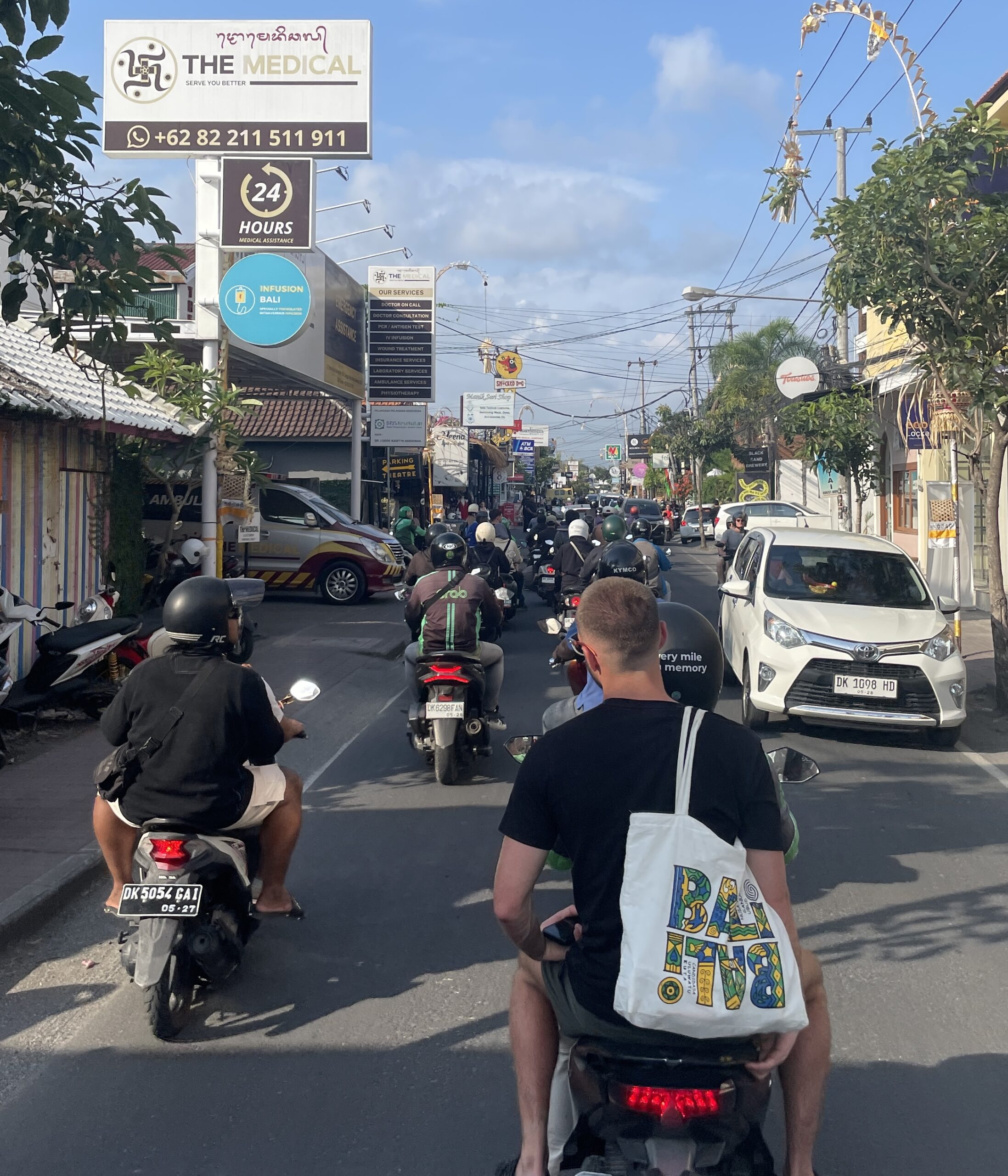 Road full of scooters in Canggu with light traffic ahead; a man rides on the back of a bike carrying a Bali tote bag.