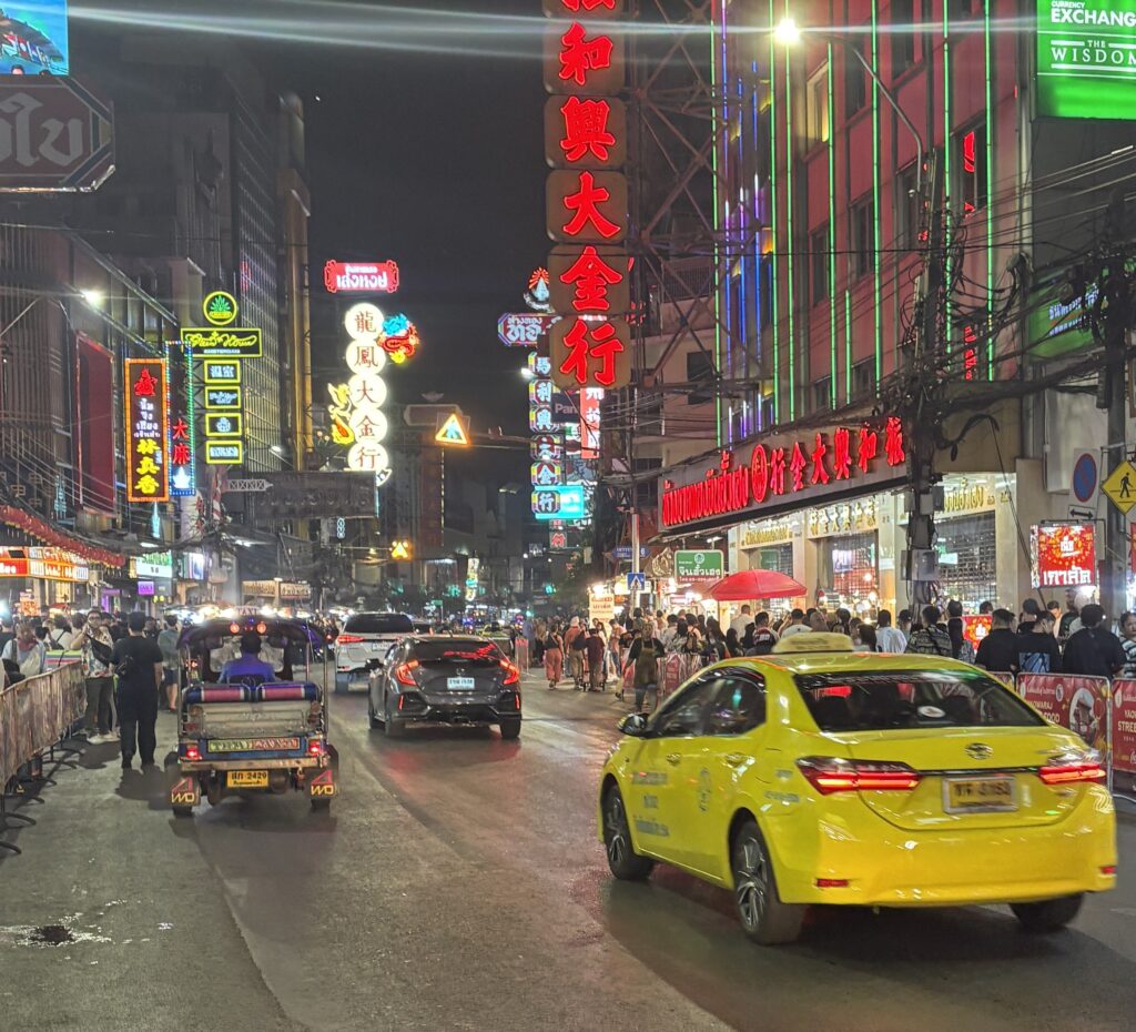 A yellow taxi drives through Bangkok’s illuminated Chinatown at night, surrounded by bright signs, locals, and tourists exploring the busy street.