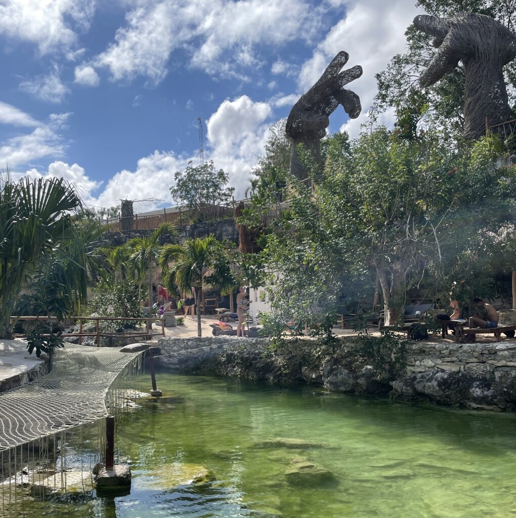 DJ performing inside Casa Tortuga cenote in Tulum with large nature sculpture and clear freshwater on a sunny day