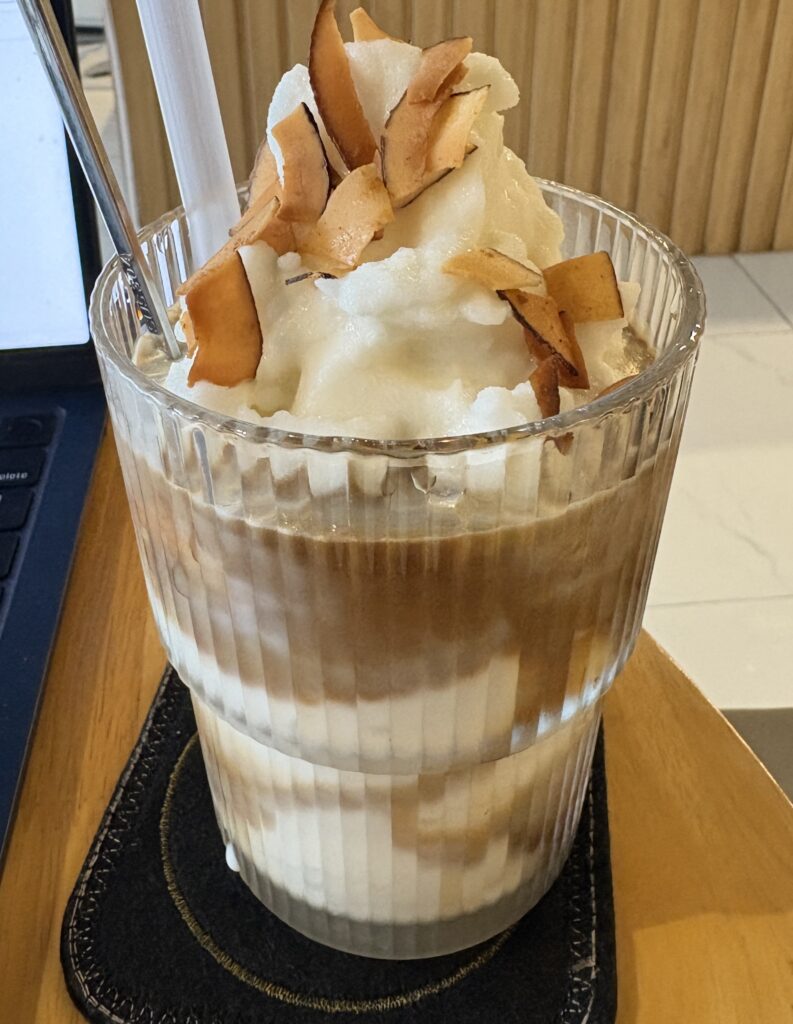 Coconut coffee topped with dried coconut chips next to a computer in a café in Da Nang
