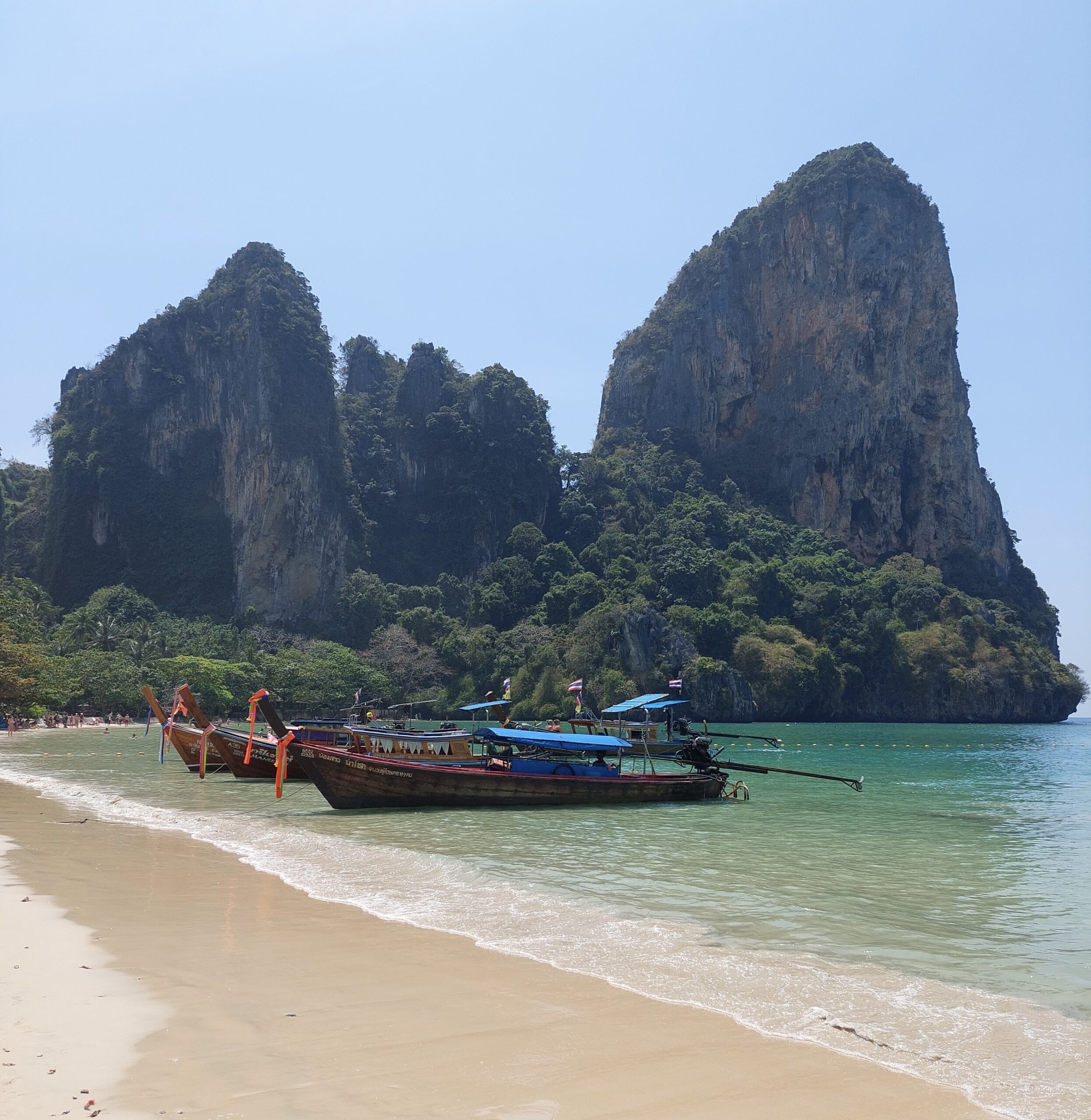 Two longtail boats on the shore of Railay Beach in Thailand with limestone cliffs and clear blue skies in the background.