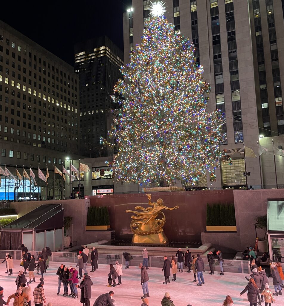 Tourists and locals ice skating at Rockefeller Center with the Christmas tree lit up at night in New York City.
