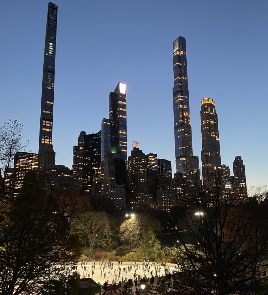 Wollman Rink in Central Park during Christmas with people skating and Billionaires’ Row skyscrapers in the background.
