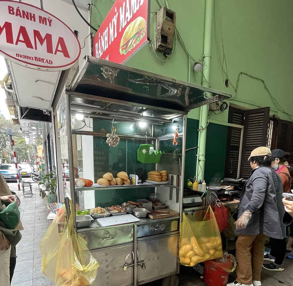 Banh Mi Mama cart in Hanoi with two women preparing banh mi sandwiches, surrounded by fresh bread and assorted toppings.