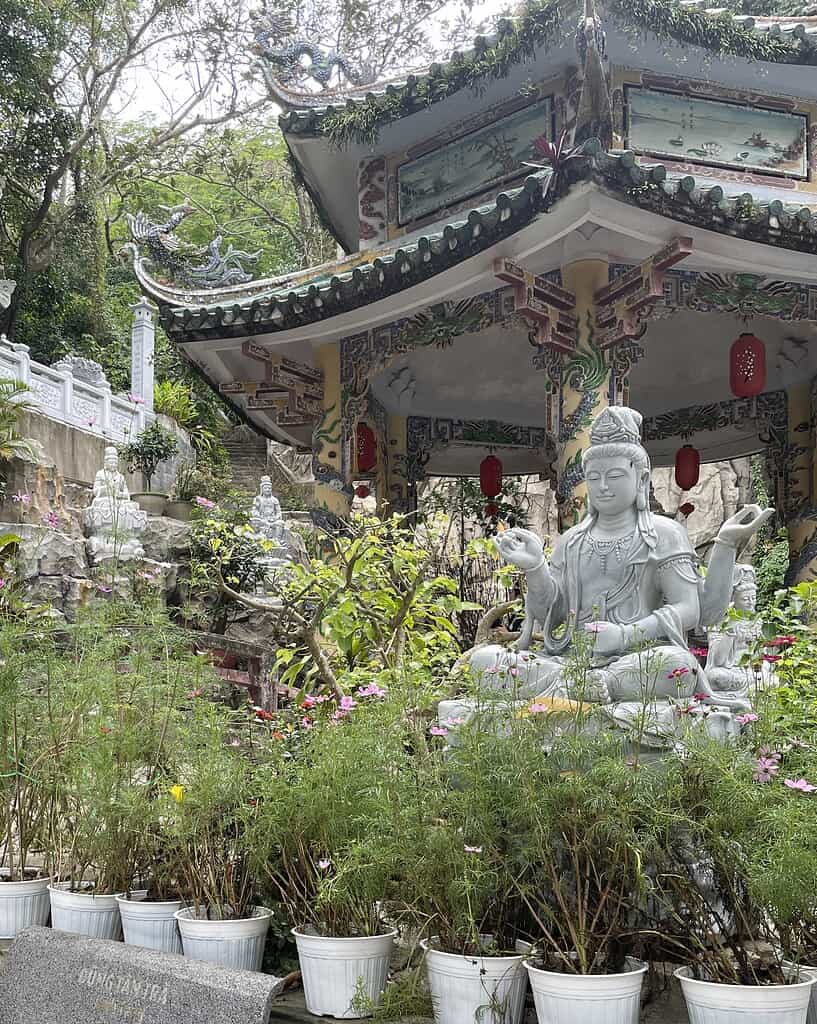 A serene Buddha statue with closed eyes and a hand gesture, surrounded by flowers and greenery, with a tall pagoda rising behind it at the Marble Mountains in Da Nang.