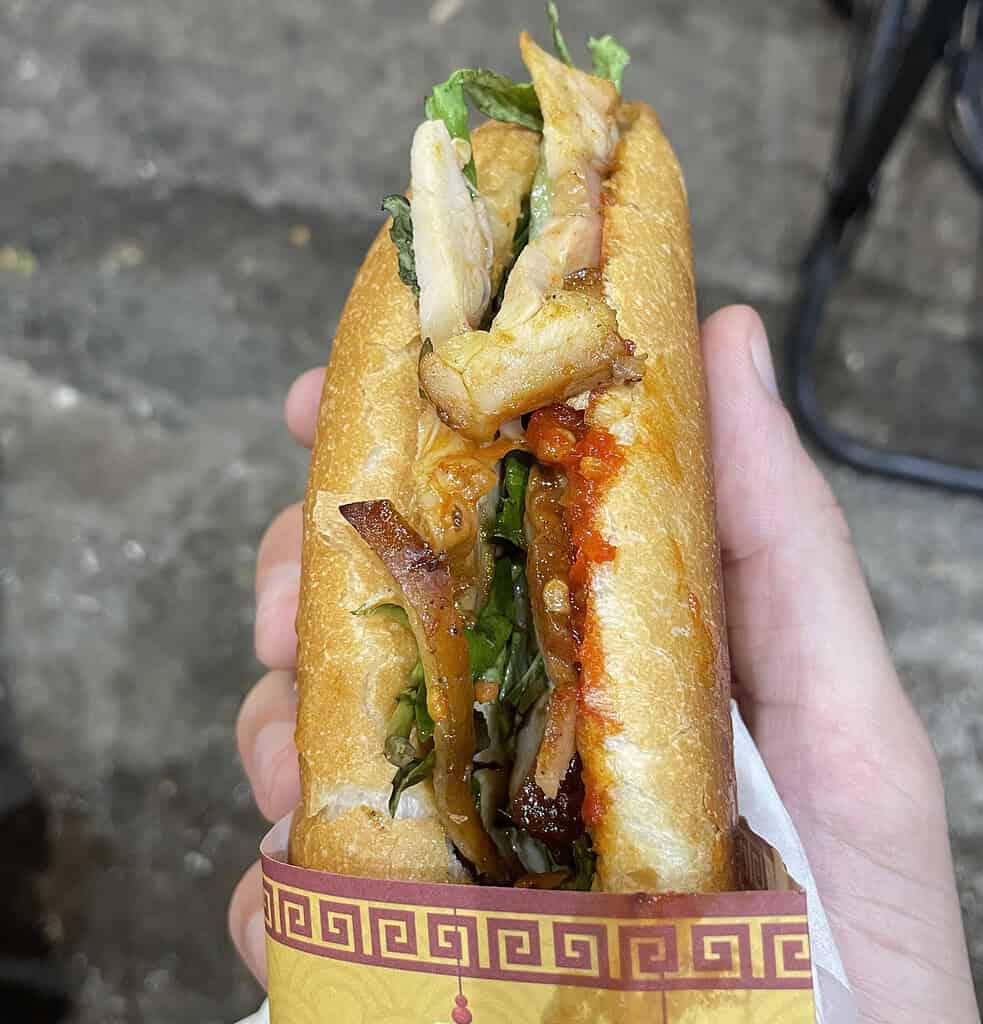 Closeup of a chicken banh mi in Hoi An, showing crispy bread, chili sauce, fresh vegetables, and sandwich wrapper underneath.