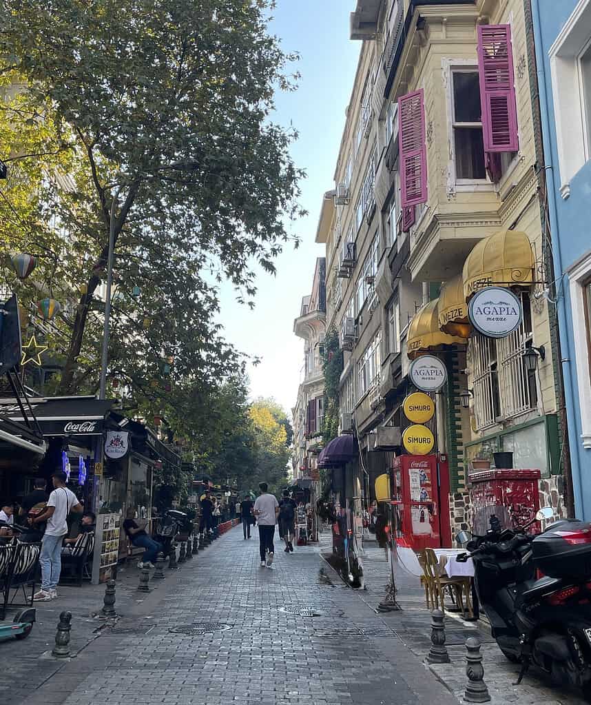 Charming cobblestone street in the Kadikoy area of Istanbul with yellow and blue old apartment buildings, a street performer, and restaurants lining both sides.