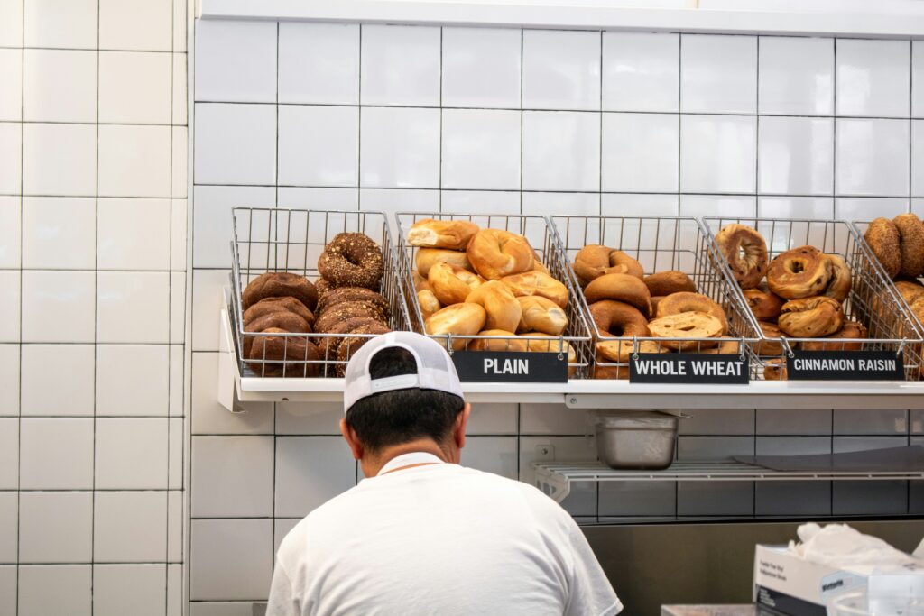 Here’s the revised alt text:

📝 Alt Text:
A man preparing a bagel behind the counter of a New York City bagel shop, with a wide variety of bagels displayed in front of him.
