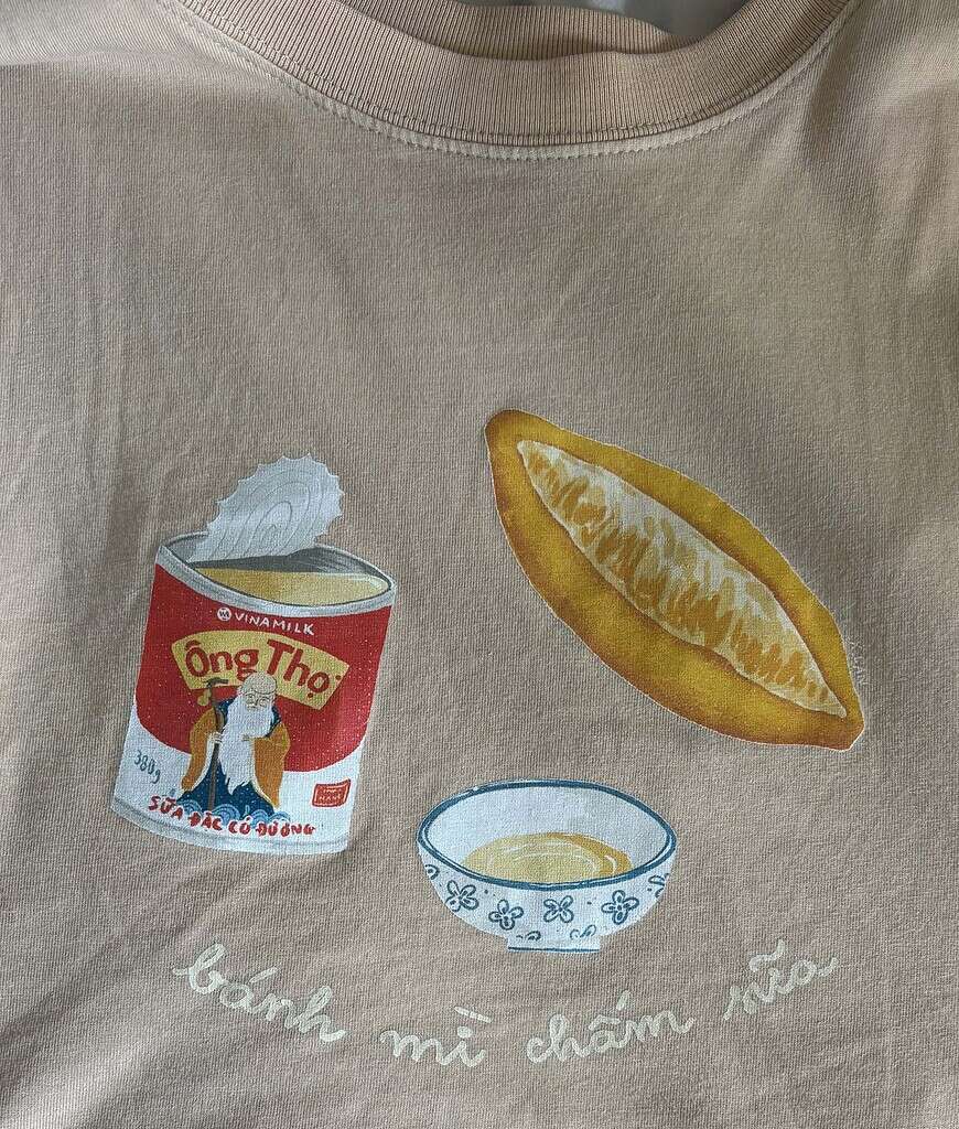 Close-up of a tan T-shirt from local Vietnamese brand TiredCity featuring a design with a bánh mì loaf and condensed milk.