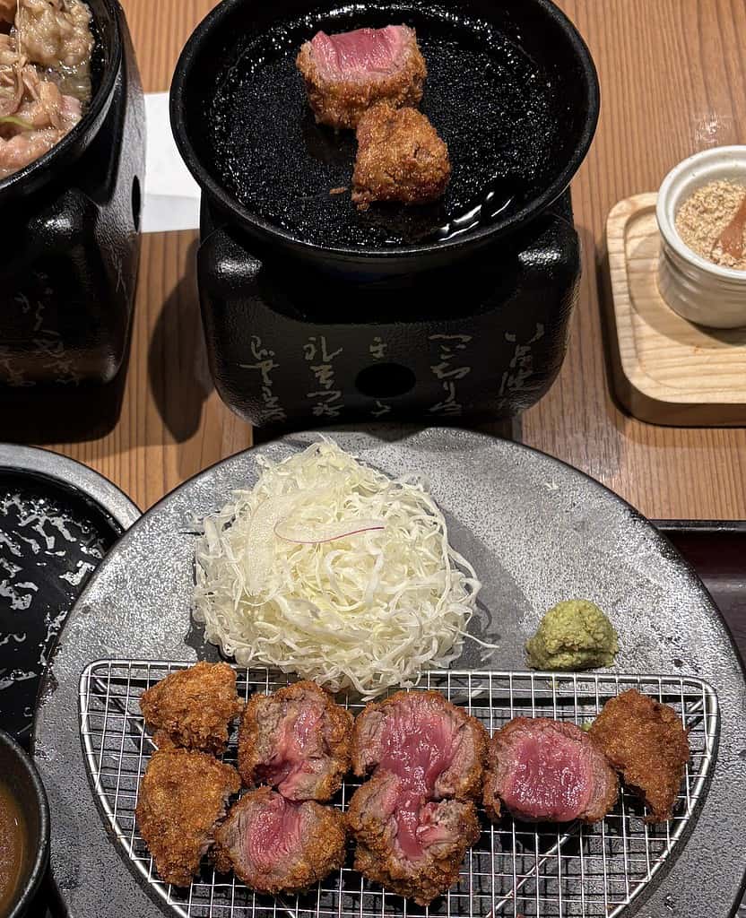 Close-up of Wagyu Gyukatsu beef cutlet with tender pink center, pieces grilling over a hot stone, served with salad and dipping sauces.