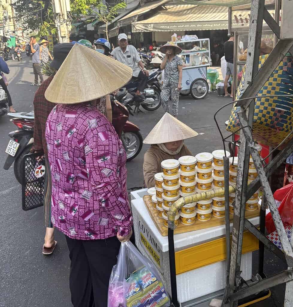 Two women wearing traditional Vietnamese conical hats shopping at a busy street market in Saigon, Vietnam