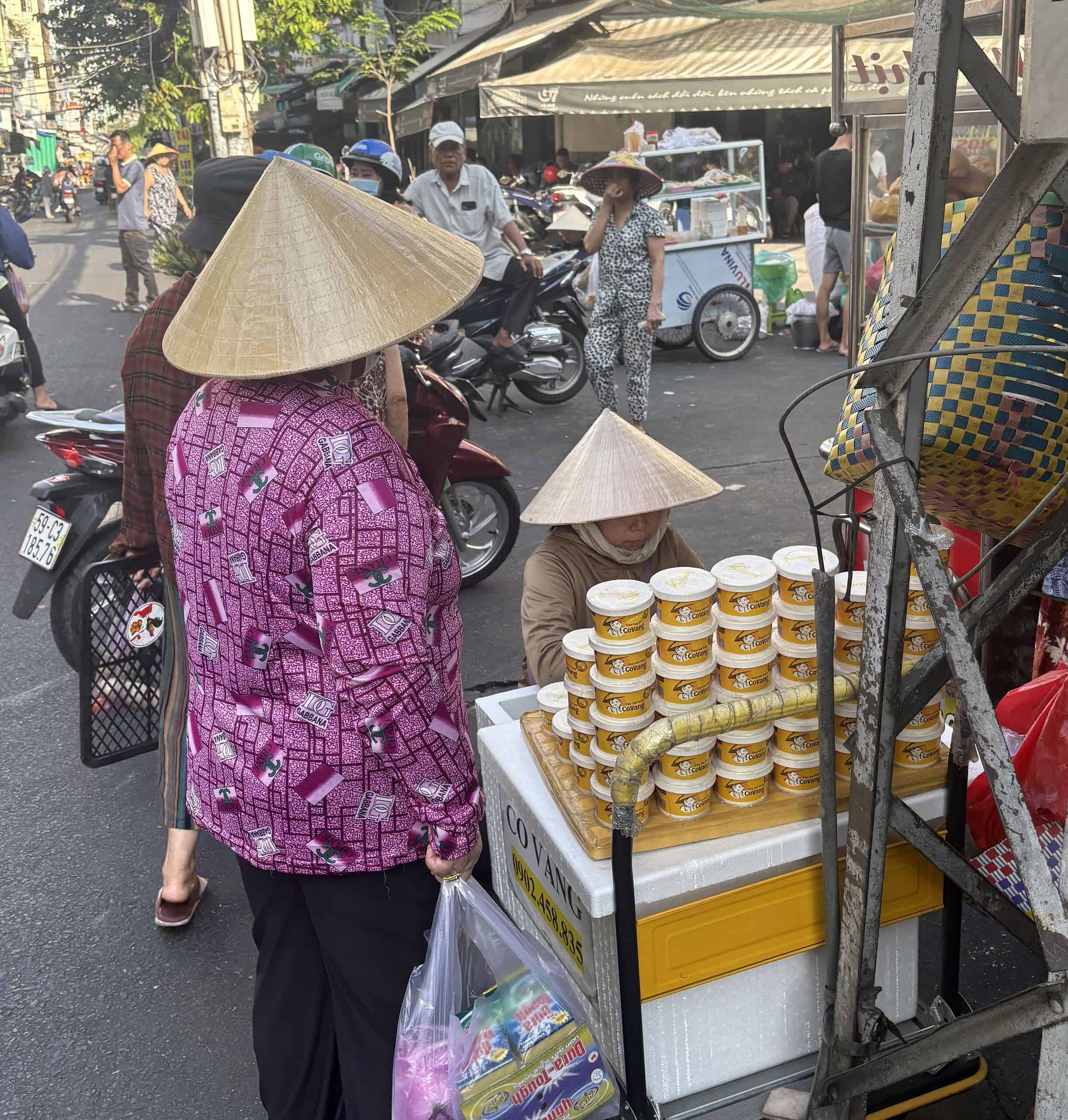 Two women wearing traditional Vietnamese conical hats shopping at a busy street market in Saigon, Vietnam