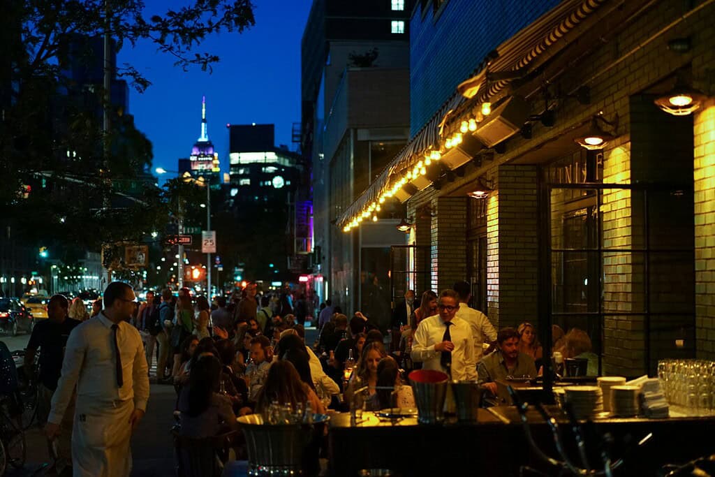 Busy street-side restaurant in New York City on a warm summer night, with people dining under string lights and the Empire State Building glowing in the background
