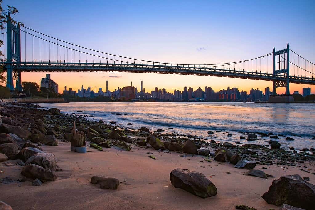 A stunning evening sunset over the East River from a small beach area in Astoria, Queens, with the sun's reflection on the water and a view of the bridge spanning the river.