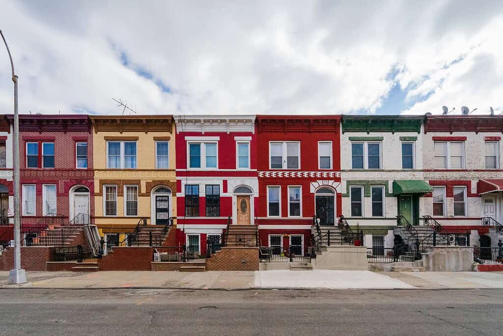  A row of iconic colorful townhouses on a street in Crown Heights, Brooklyn, including a prominent bright yellow home under a gray, cloudy sky.