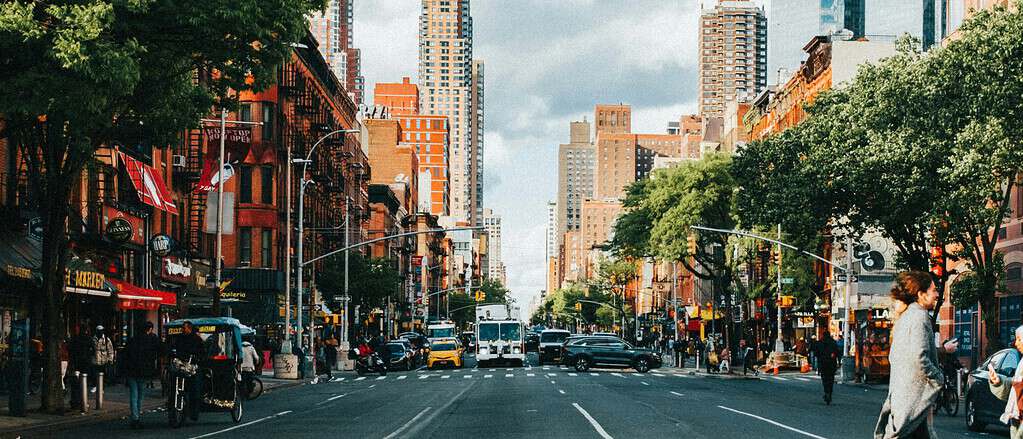 A lively street scene in Hell's Kitchen during early September, showing oncoming traffic and city lights at dusk from a crosswalk perspective with autumn energy in the air.