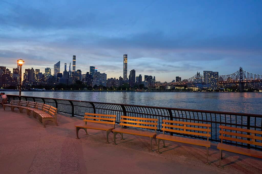 A wide sunset view of the Manhattan skyline across the East River from Long Island City, featuring deep orange and purple skies behind iconic skyscrapers.
