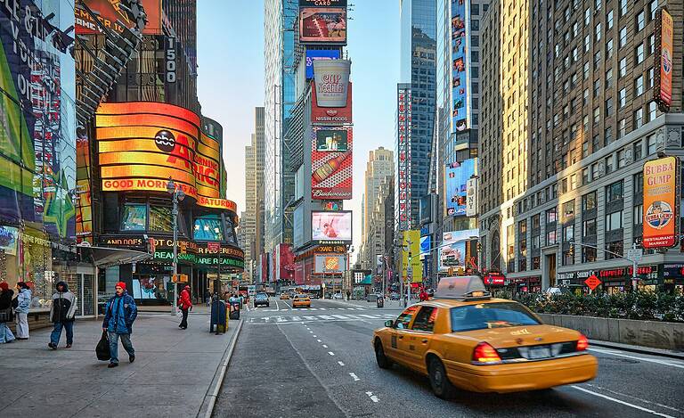 Times Square during the day with yellow taxis, bright billboard ads, and pedestrians walking through Midtown Manhattan