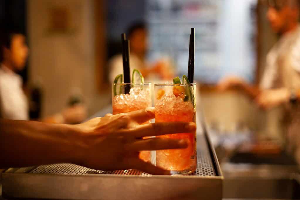 A bartender’s hands holding two vibrant mixed cocktails over a dark bar counter, signaling the start of happy hour in a New York City lounge.
