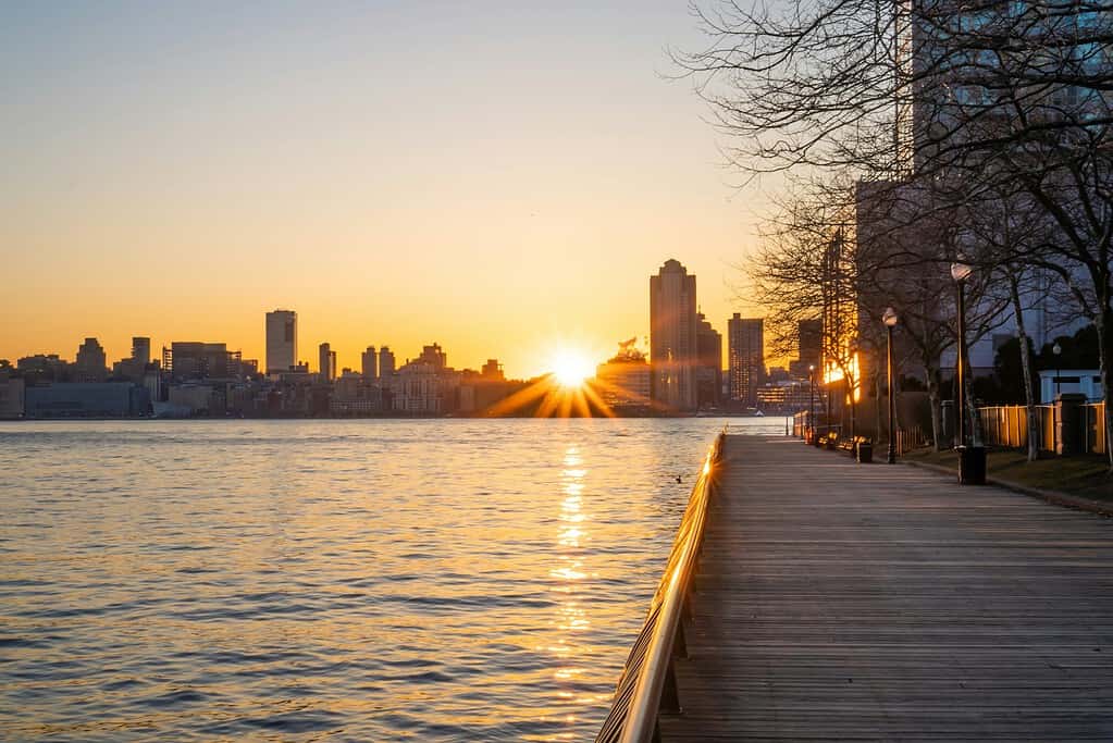 A scenic view of the Hudson River at sunset from the Jersey City waterfront, featuring the glowing New York City skyline across the water under a vibrant sky.