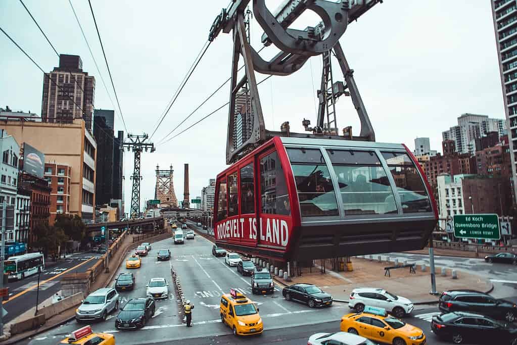 A red Roosevelt Island Tram cabin in motion, suspended in the air above the East River and alongside the Queensboro Bridge, with the Manhattan skyline and Roosevelt Island in the background.
